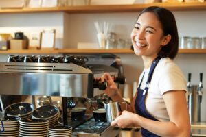 Happy asian girl barista, working in cafe, brewing coffee, prepare cappuccino, using steamer on